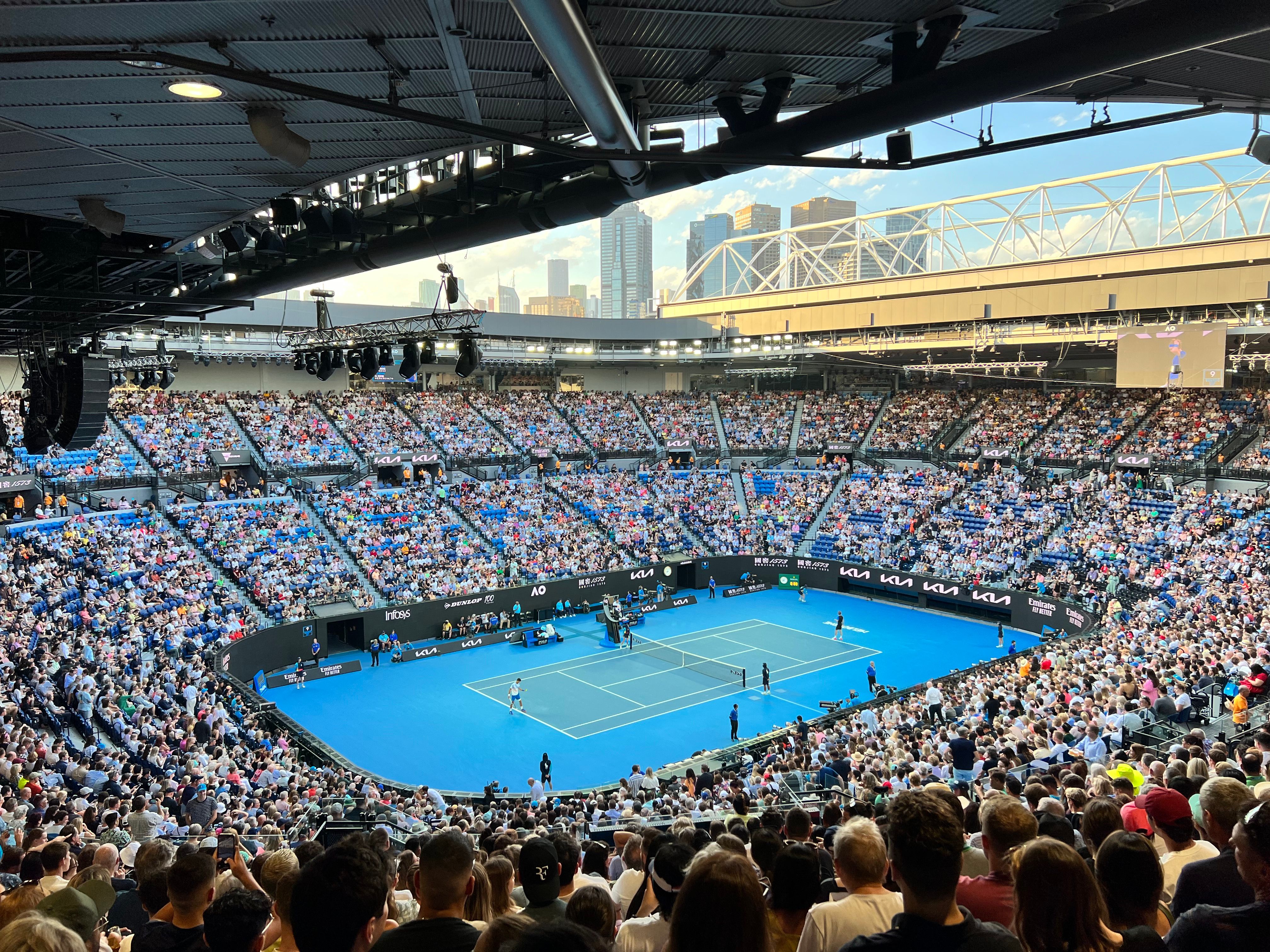 A full Rod Laver Arena watching a match on the blue hard court, with the city skyline beyond the open roof