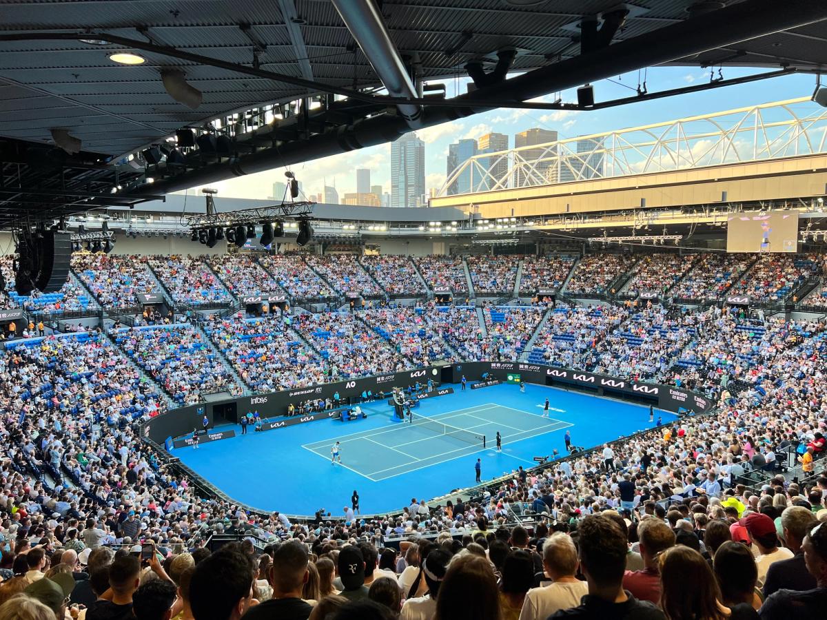 A full Rod Laver Arena watching a match on the blue hard court, with the city skyline beyond the open roof