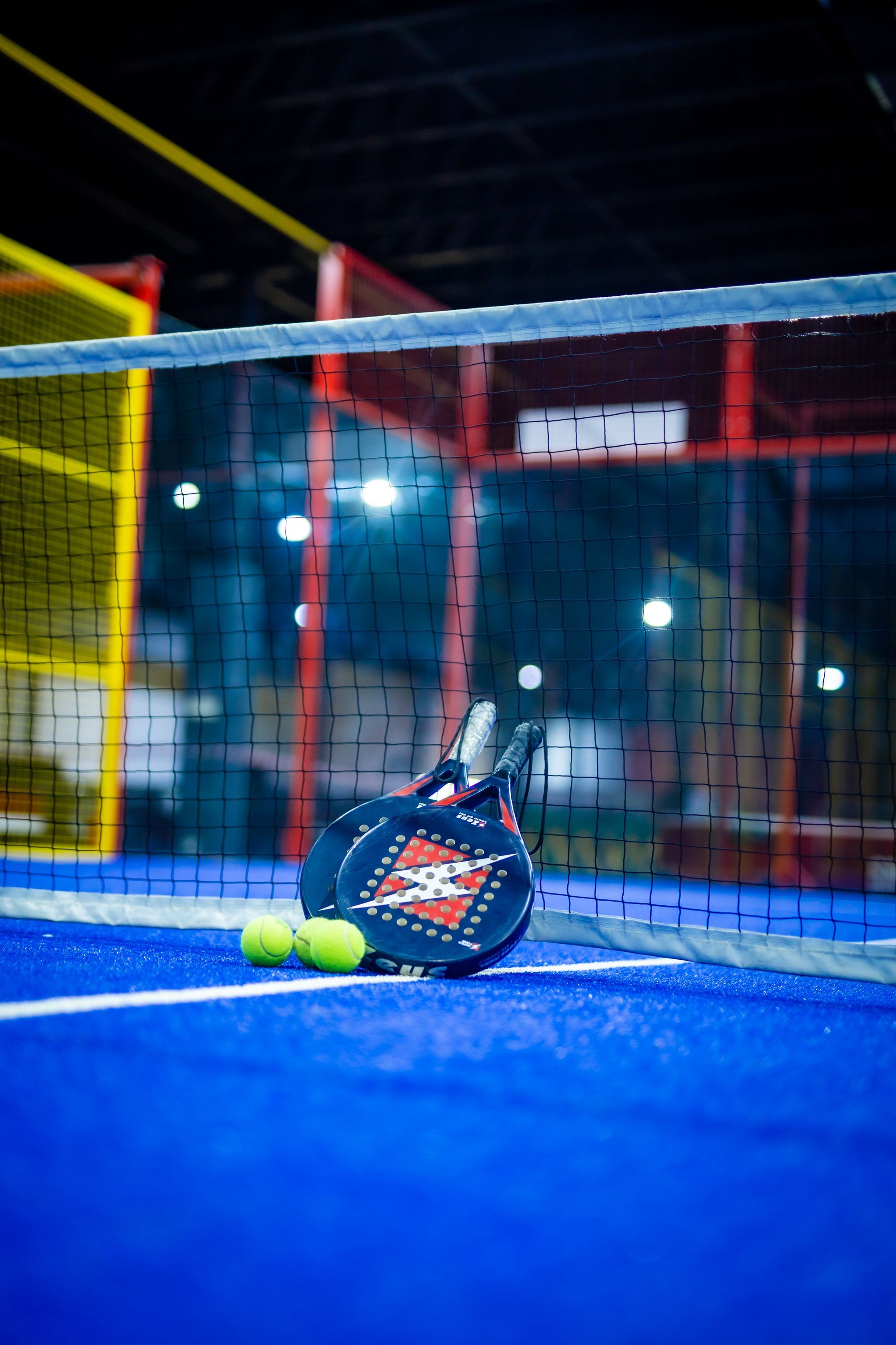 Two padel rackets and two yellow balls resting against the net on a blue indoor padel court, with mesh fencing and floodlights behind