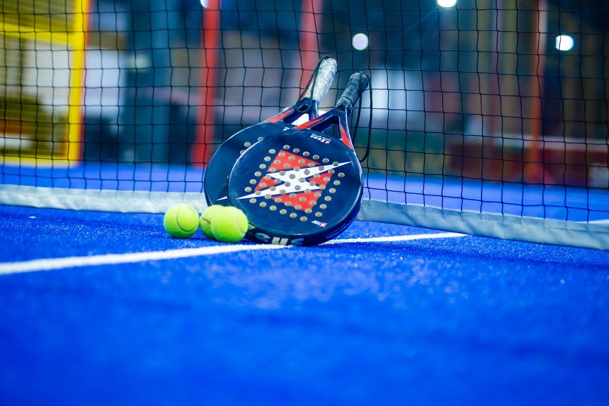 Two padel rackets and two yellow balls resting against the net on a blue indoor padel court, with mesh fencing and floodlights behind
