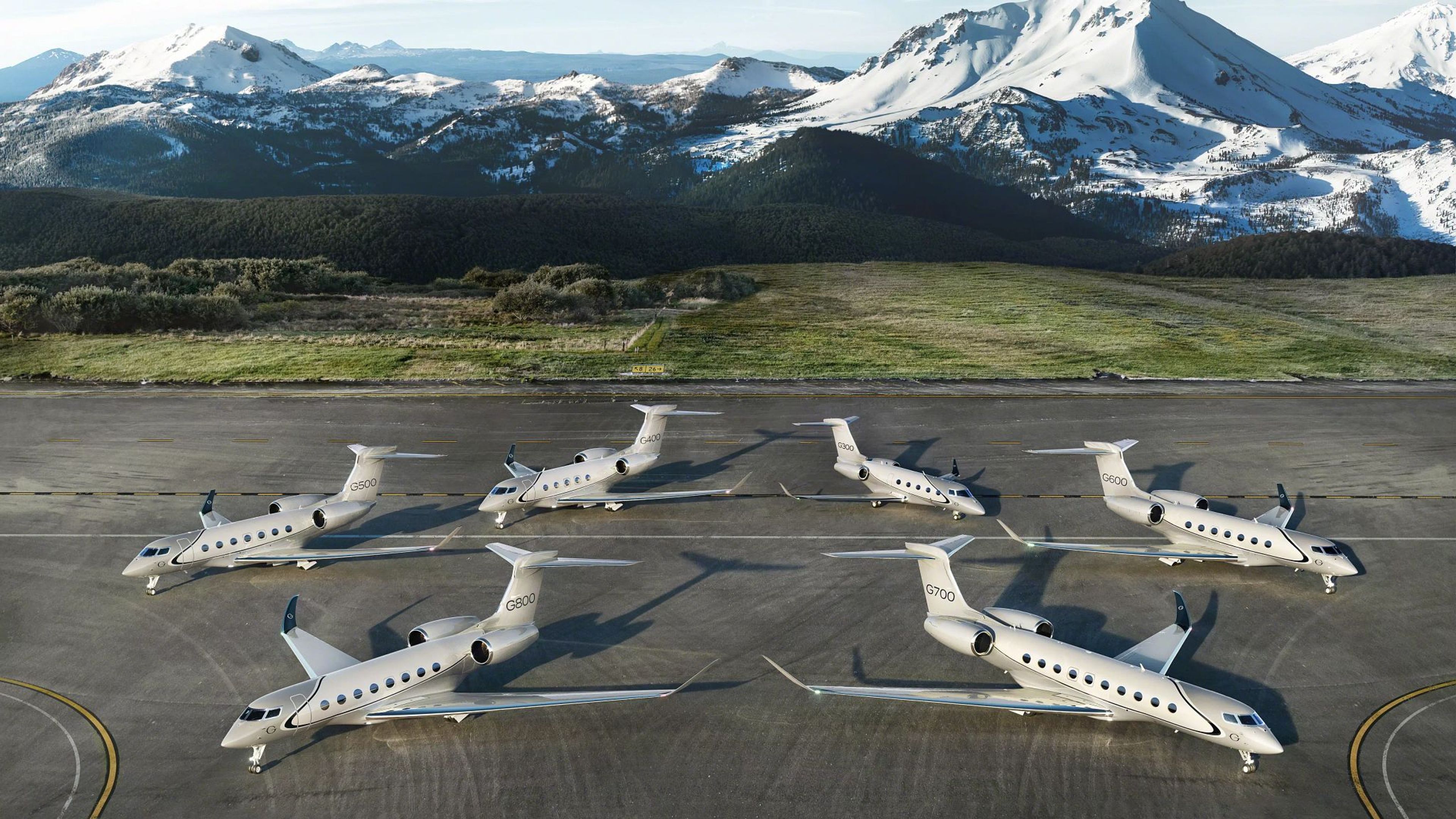 Alt Text Six Gulfstream private jets arranged on an airfield apron facing in opposite directions, with snow-capped mountain peaks in the background under a clear sky.