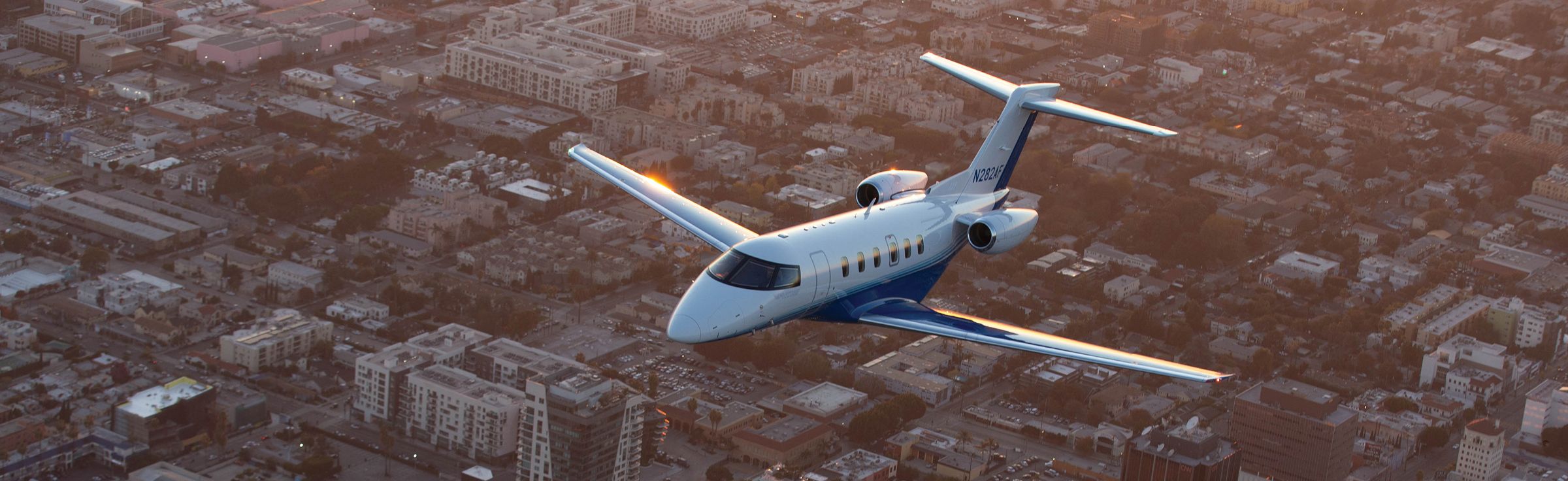 PlaneSense Pilatus PC-24 jet flying over a city in warm evening light.