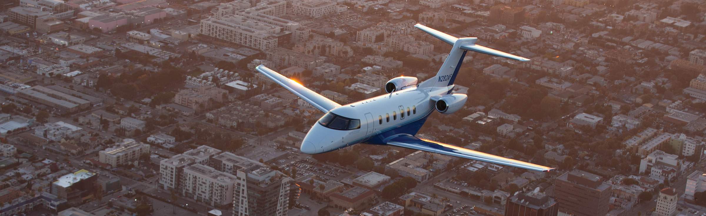 PlaneSense Pilatus PC-24 jet flying over a city in warm evening light.