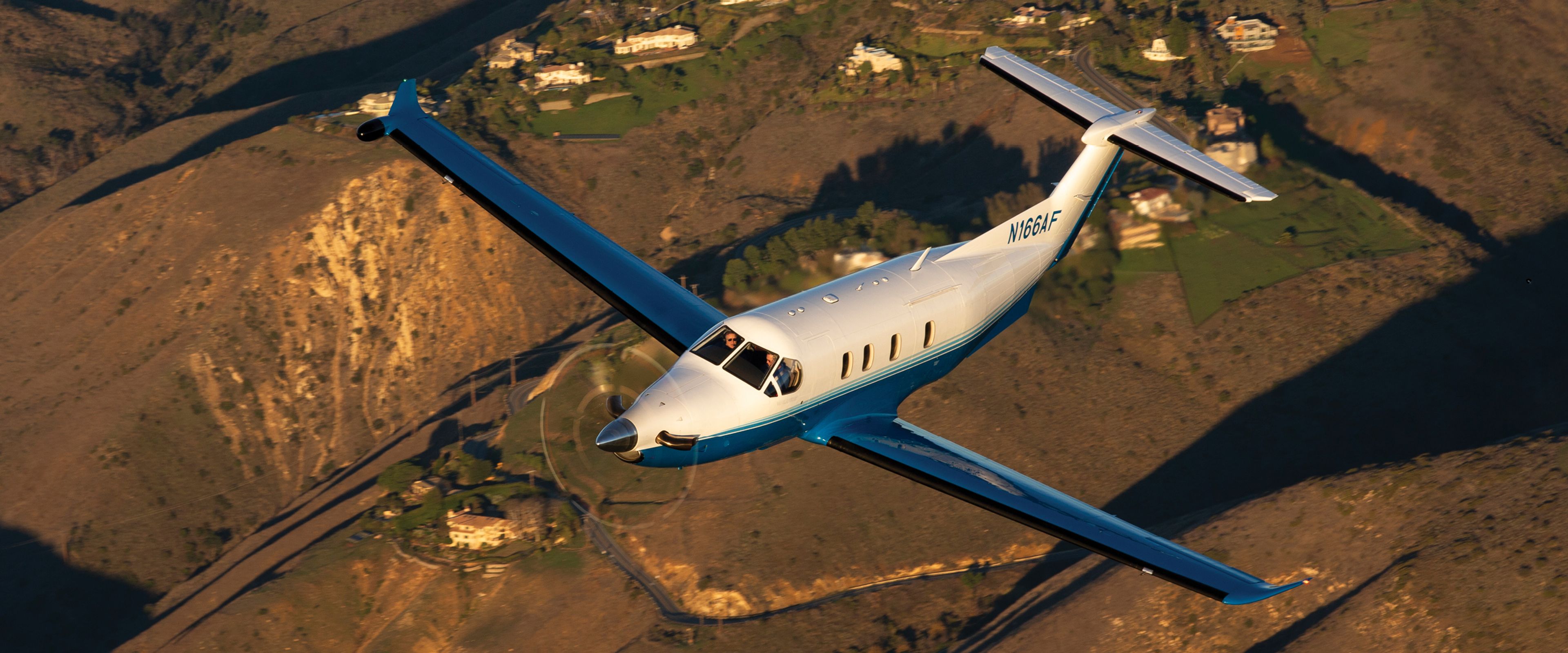 PlaneSense Pilatus PC-12 turboprop flying over hills in blue and white livery.