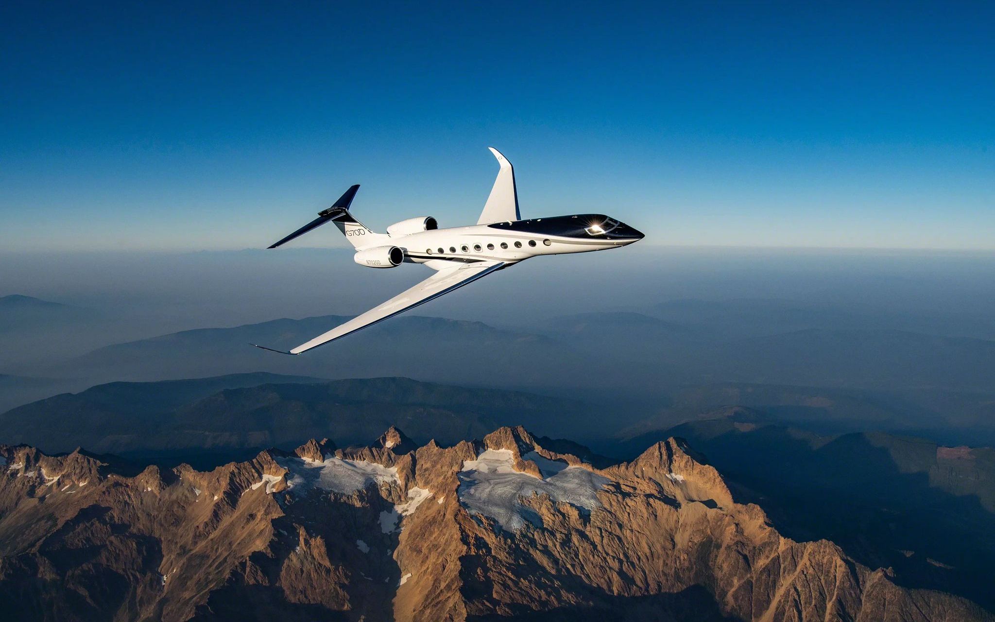 White Gulfstream G700 banking above a rugged, snow-streaked mountain range beneath a clear blue sky.