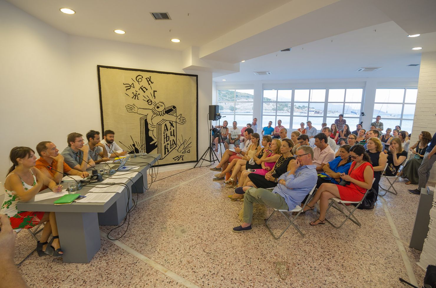 Public in Art Space Pythagorion's main hall seating on chairs in front of the hall's window with a view to the sea looking at a group of speakers seating at a long grey table.