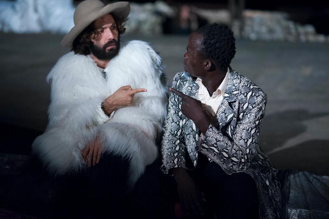 Two men sitting next to each other, looking and pointing a finger at each other