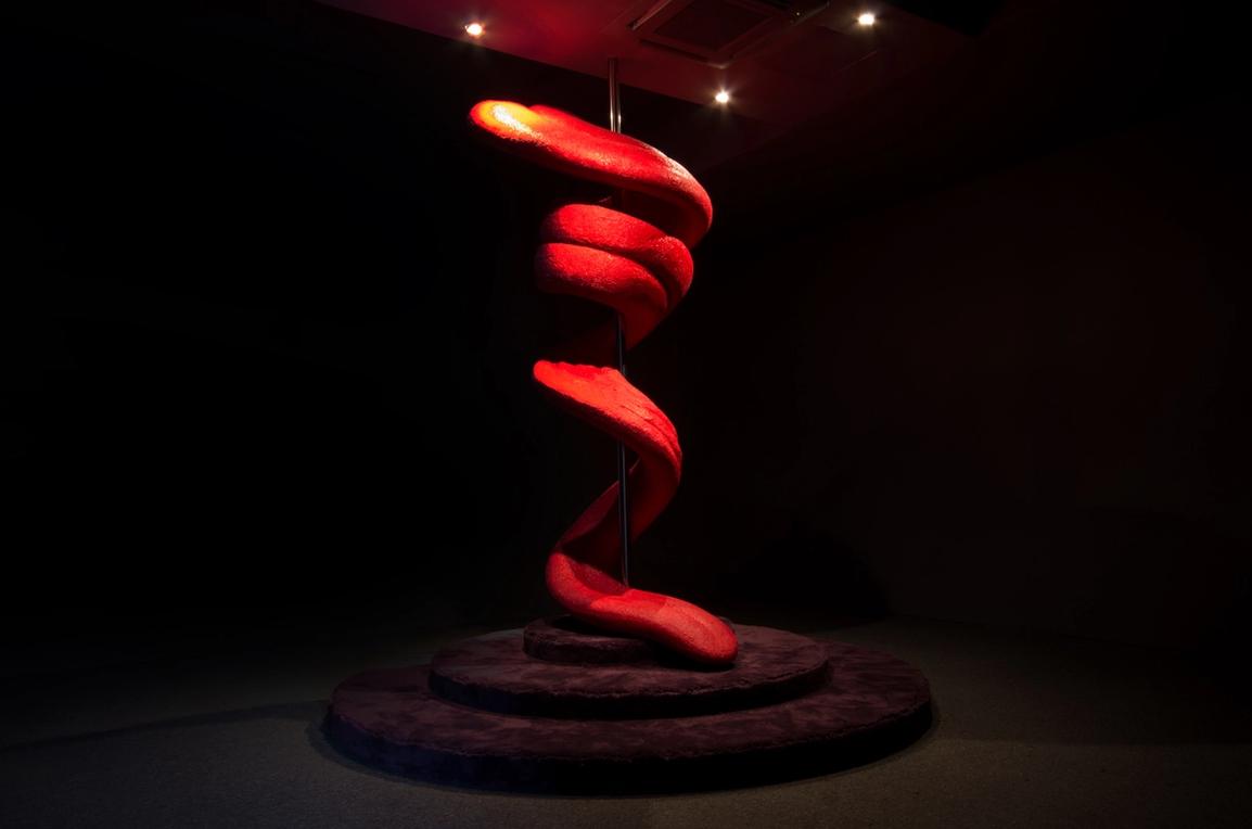 A giant sculpture of a fleshy tongue twirling around and down a metal dancing pole in a dark room.