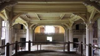 A picture inside of a ruin. The viewer ist on a wooden podium, everything is covered in dust, the big room is fully build out of wood with classicist decorated columns. The viewer looks toward a stage.