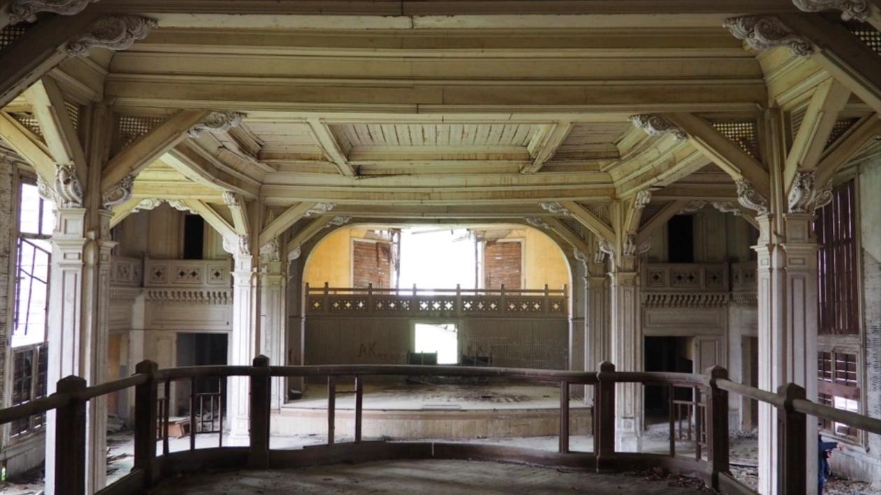 A picture inside of a ruin. The viewer ist on a wooden podium, everything is covered in dust, the big room is fully build out of wood with classicist decorated columns. The viewer looks toward a stage.
