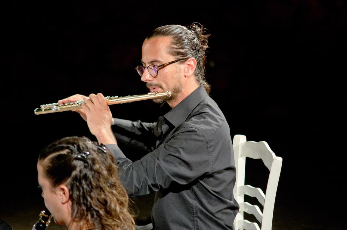 Mehmet Can Aglac playing the flute. He's wearing a black shirt, horn glasses and long tied back hair. In the front you can see the back of the head of the clarinettist, Yonca Alpay