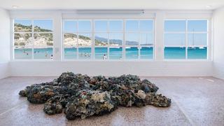 Das Werk von Maarten Vanden Eynde, eine Skulptur aus Plastikmüll, der aus den fünf großen Meereswirbeln stammt, mit dem Titel "Plastic Reef", befindet sich im Vordergrund der Haupthalle des Art Space Pythagorion. Im Hintergrund blickt das große Fenster der Halle auf das blaue Meer, die Schwimmer und die Küsten der Türkei.