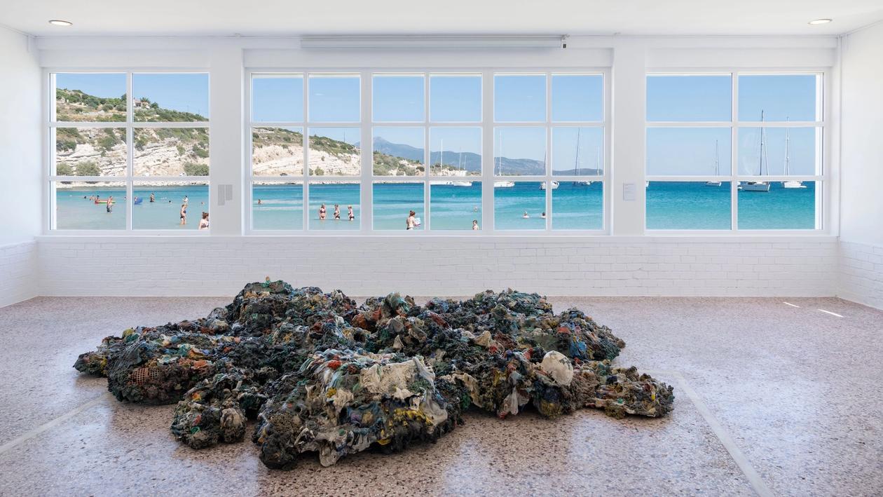 Maarten Vanden Eynde's work, a sculpture made of plastic debris coming from the five major oceanic gyres, titled "Plastic Reef" is situated in the foreground in the main hall of Art Space Pythagorion. In the background, the hall's large window faces the blue sea, swimmers and the shores of Turkey.
