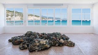 Maarten Vanden Eynde's work, a sculpture made of plastic debris coming from the five major oceanic gyres, titled "Plastic Reef" is situated in the foreground in the main hall of Art Space Pythagorion. In the background, the hall's large window faces the blue sea, swimmers and the shores of Turkey.