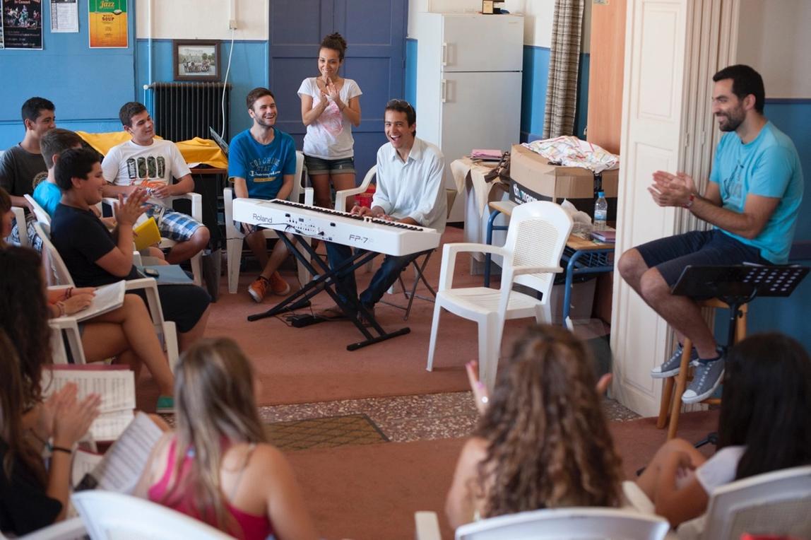 Guy Mintus giving a class to Samian Musicians. He's sitting at a Keyboard with a bright smile, while he's surrounded by pupil looking at him, clapping and smiling