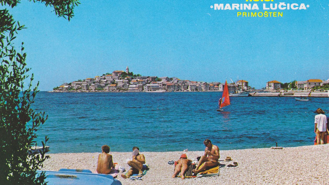 A 70s period postcard showing a nudist beach in Crotia with people subathing and swimming. In the background is a peninsula with houses