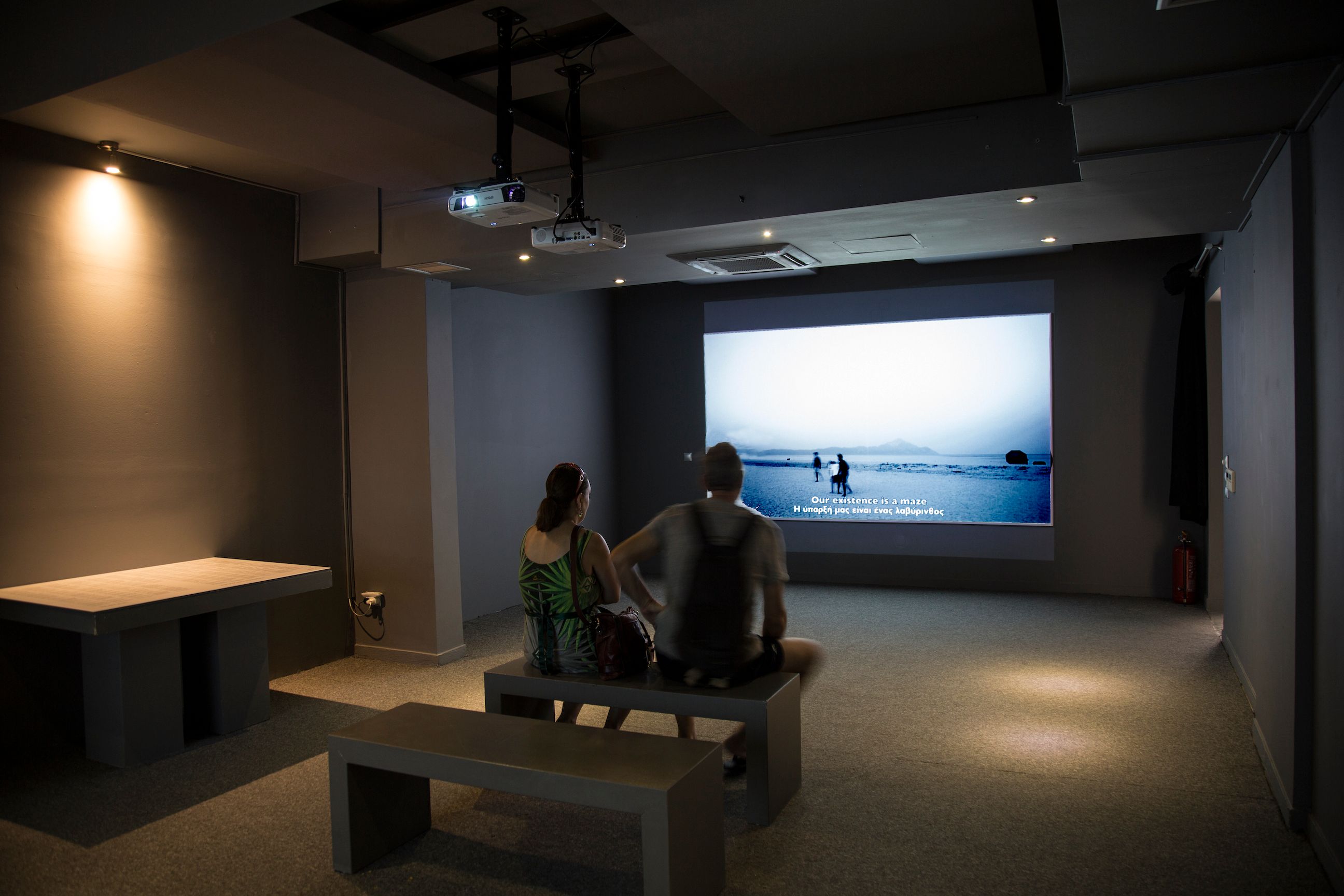 Two people seating on a bench in a projection room watching a video by Nicolas Kozakis & Raoul Vaneigem