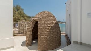 Ένα στρογγυλό κτίσμα με ψηλό θόλο φτιαγμένο άπο πλίνθους με μια μικρή είσοδο έχει κατασκευαστεί στον υπαίθριο χώρο του Art Space Pythagorion. Στο βάθος φαίνεται η παραλία και η θάλασσα.
