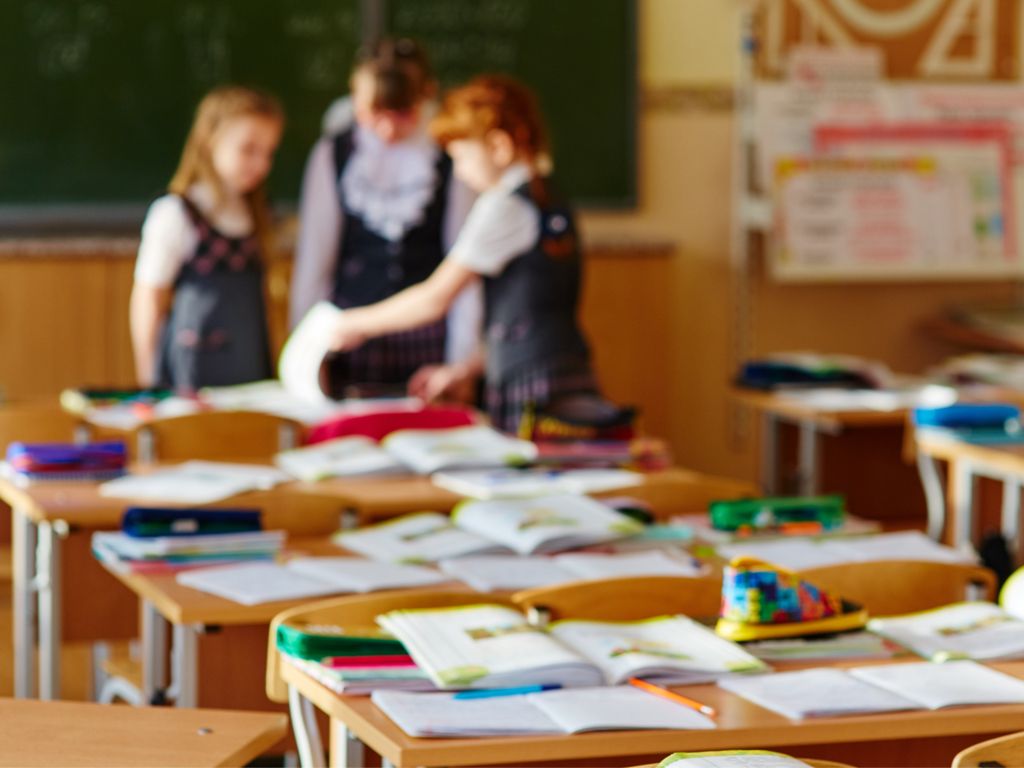 A class room with tables full of open books, pens and leaflets. In the background, blurry, three pupils talking, standing around a table.