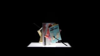 Μια παραδοσιακή σούβλα κεμπάπ διαπερνά βιβλία και έντυπα. Το έργο βρίσκεται πάνω σε μια φωτεινή τετράγωνη επιφάνεια σε μαύρο φόντο.