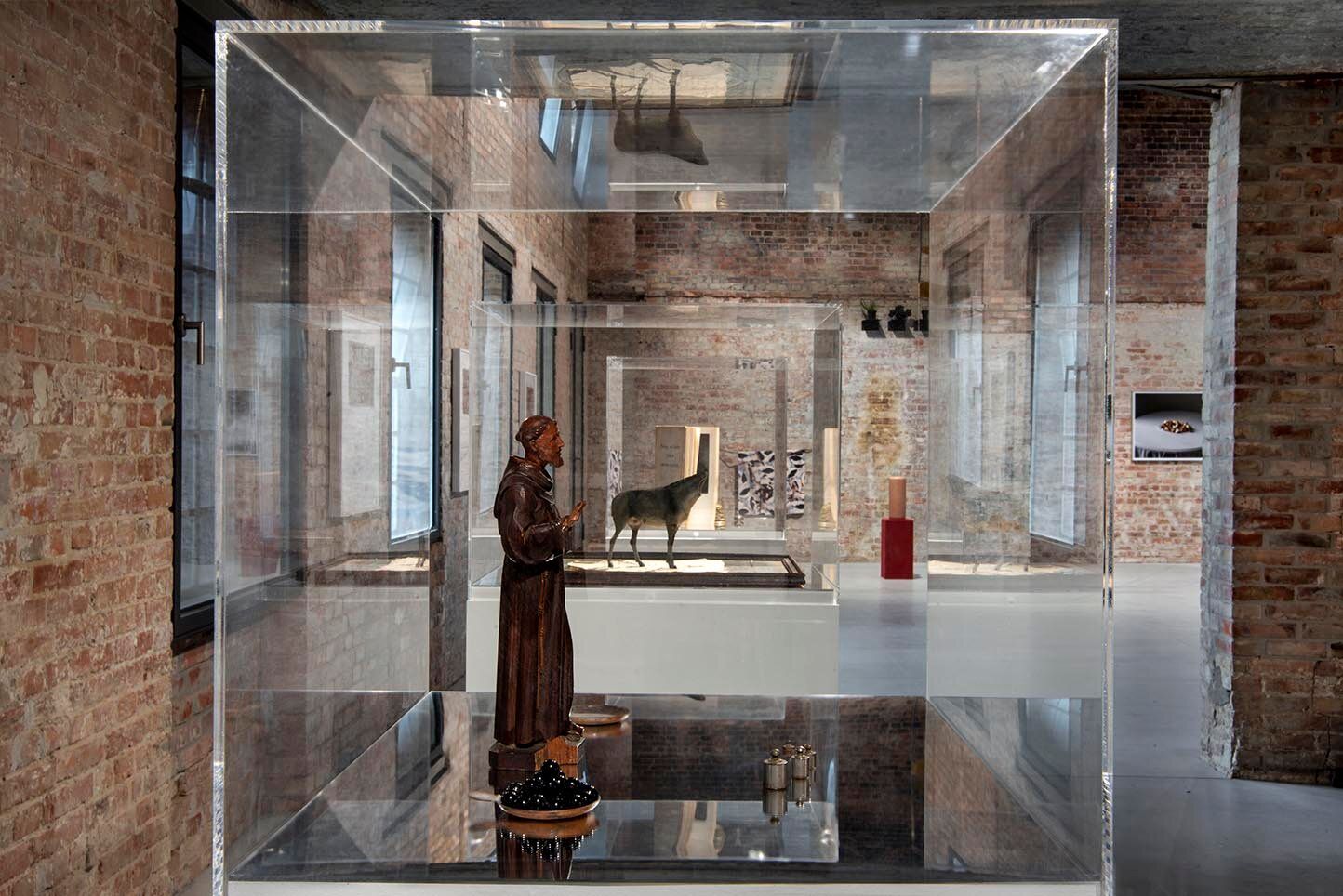 A human figurine of a catholic monkwith smaller items place around him showcased in a gallery space with walls made of bricks.