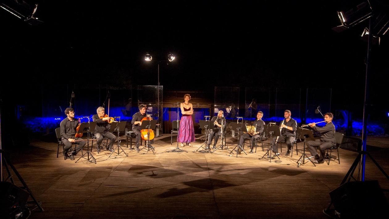 Mezzo-soprano Lenia Safiropoulou standing in the middle of the illuminated stage of the Samos Young Artists Festival in a purple dress. On her left seated the viola, violin and cello. On her right seated, the flute, clarinet, french horn and oboe