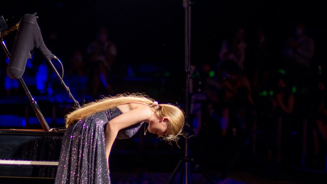 Eine junge Musikerin in einem glitzernden dunklen langen Kleid mit langem blonden Haar macht eine tiefe Verneigung auf der hell beleuchteten Bühne des Antiken Theaters von Pythagorion, bei Nacht 