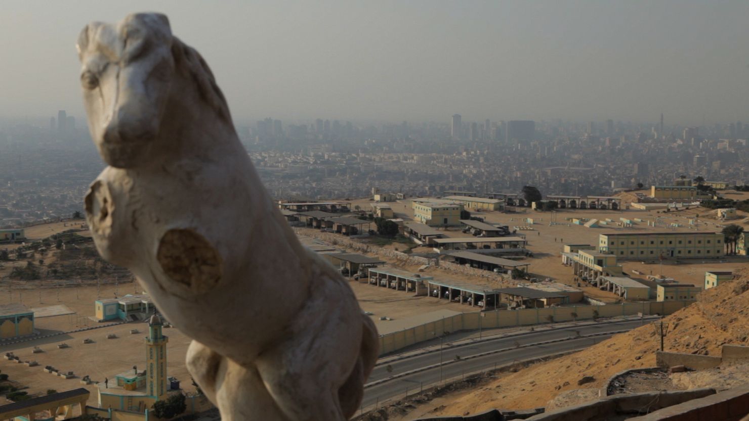 A marble sculpture of a horse-like animal with mutilated upper limbs in front of an area with industrial-looking infrastructure and. A city of the middle east can be seen in the background.