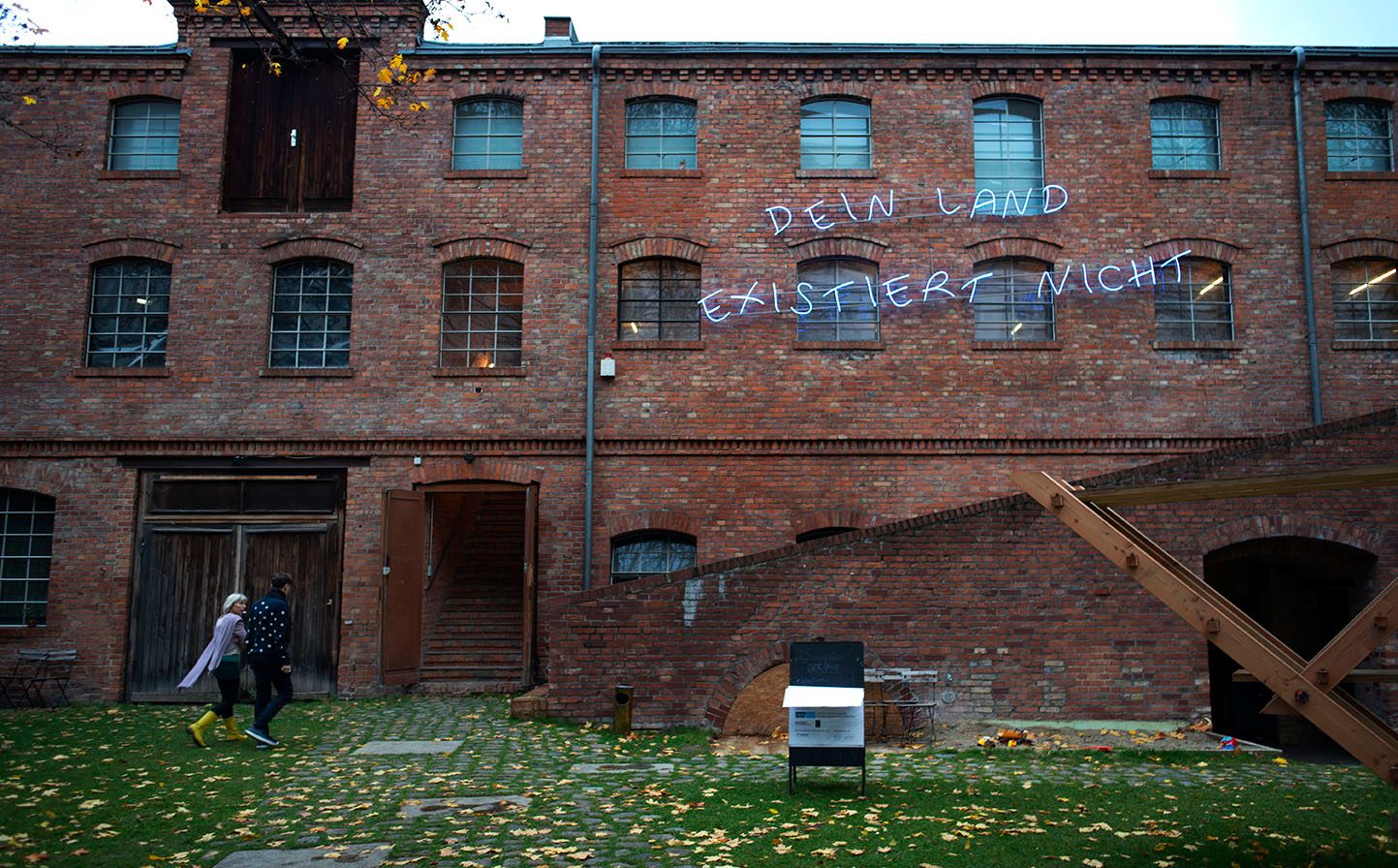 Stephan Koal and Katja Ehrhardt, walking, in front of Freiraum in der Box Exhibition Space. The building is made out of red bricks and features on its facade the phrase: "Dein Land Existiert Nicht" in neon letters. Green grass and falled yellow leaves are in front of the building.