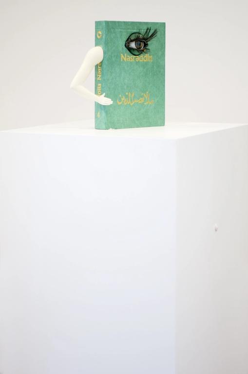 A green book is placed on a white pedestal in white interiors. The book has a plastic human arm and an eye attached on its cover. Its title, in golden letters, is written in arabic.