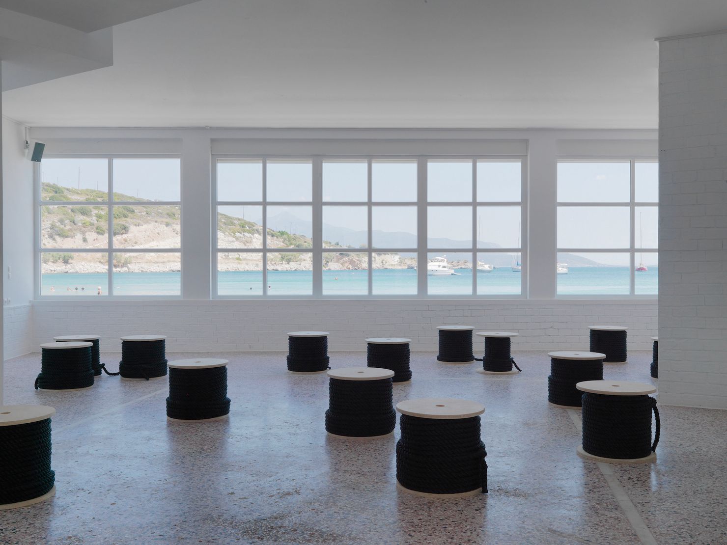 Art Space Pythagorion's main hall with a view to the open sea and the shores of Turkey. On the hall's red terrazzo floor, dark fishing rope is wrapped around fifteen wooden coils distributed in the space.