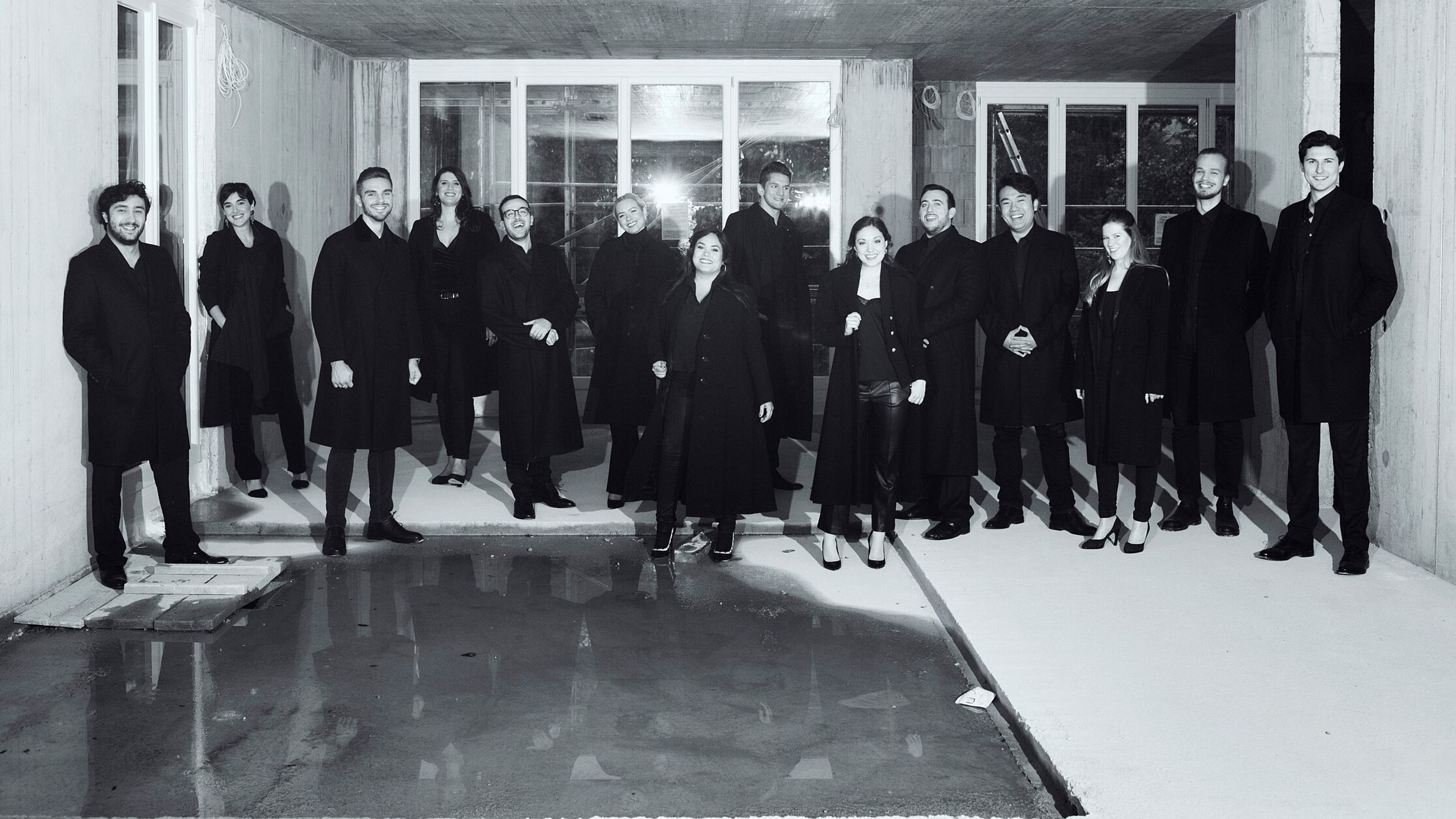 Ein Schwarz-Weiß-Foto von 14 Studenten in Mänteln. Alle lächeln in die Kamera und stehen nebeneinander, wie für ein Gruppenfoto, aber nicht in einer Reihe und mit einem Abstand zwischen ihnen.