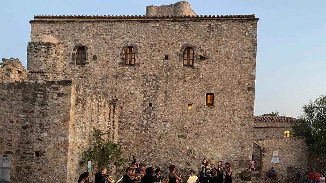 A group of musicians in front of the ruins of a Greek castle in a summer evening light. At the border of the image you can see a row of visitors of the concerts on plastic chairs