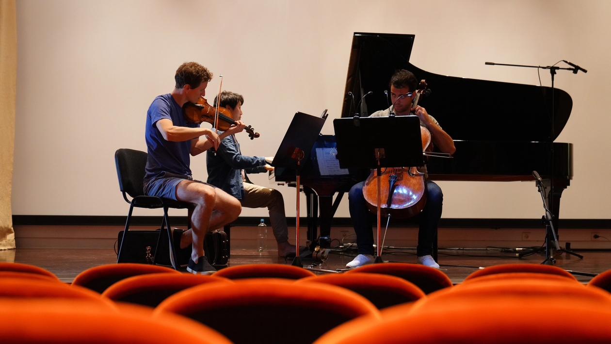 All three musicians with everyday clothes rehearsing on stage, each one playing their music instrument. The hall's empty seats are out of focus.