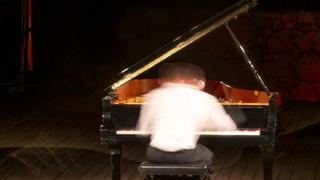 Picture of a piano player playing the piano on stage. He wear a white shirt and dark pants. The picture is taken with long exposure