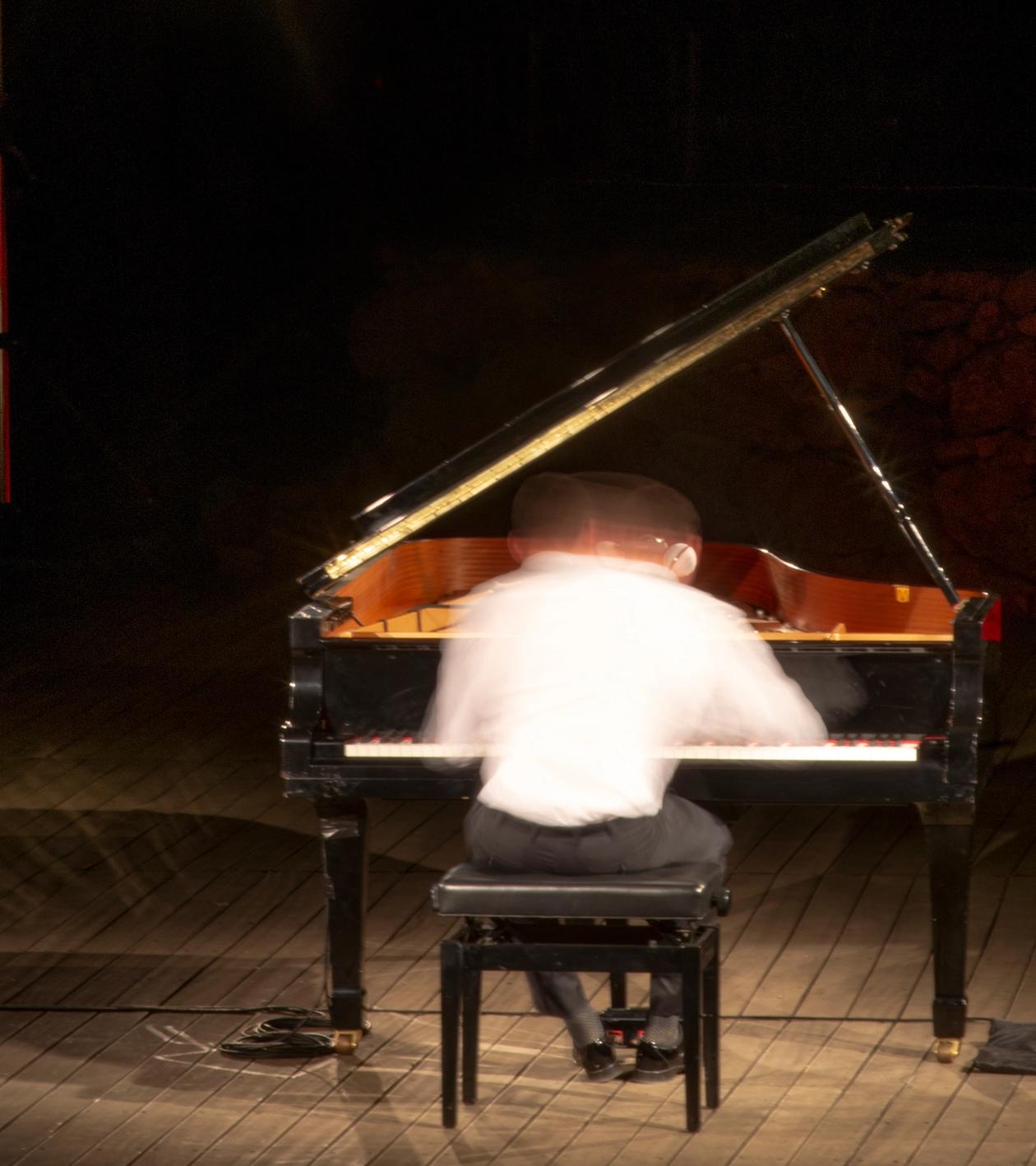 Picture of a piano player playing the piano on stage. He wear a white shirt and dark pants. The picture is taken with long exposure