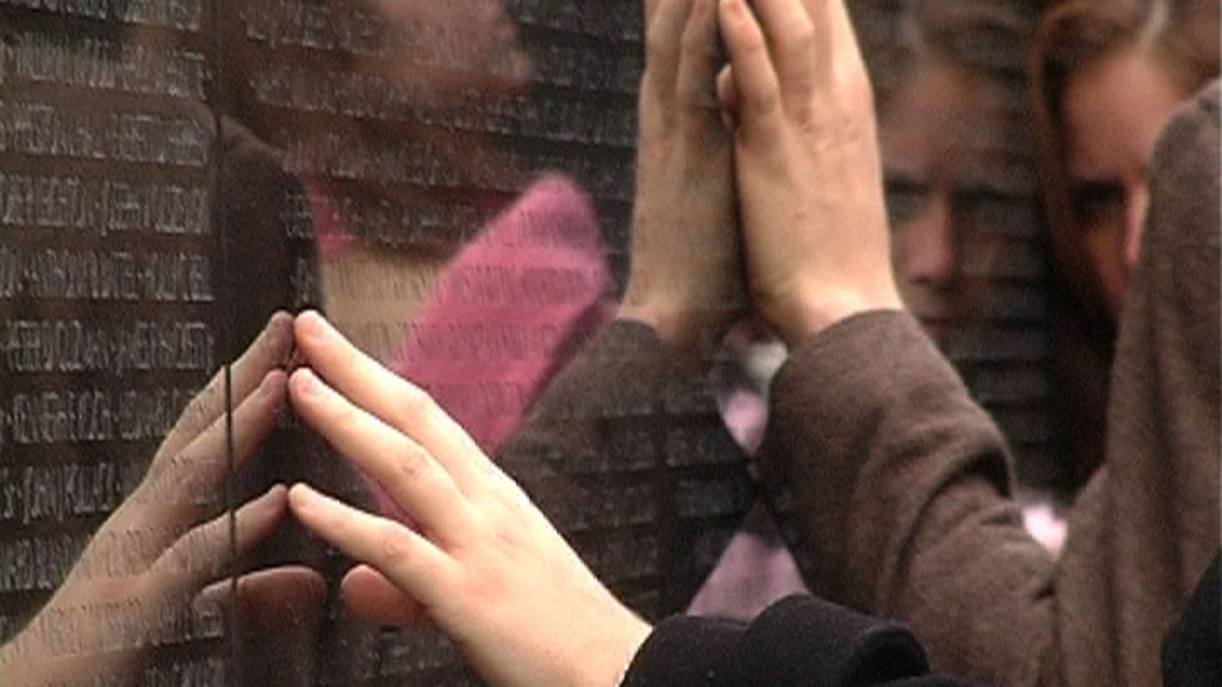 Video still of a close up shot of hands touching a black surface with engraved letters. The surface is polished and the reflection of some people's faces can be seen.