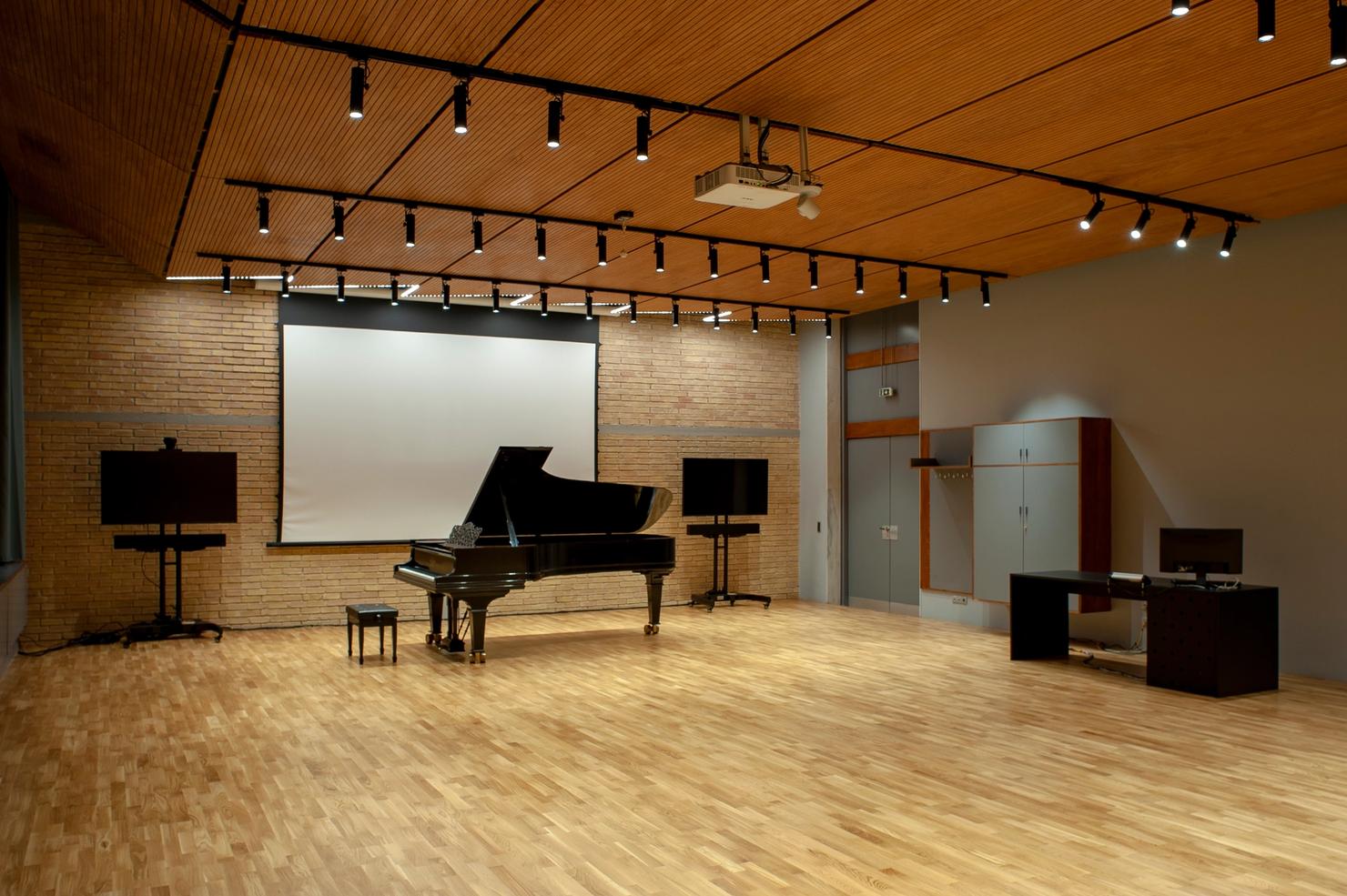 The distance learning studio is a room within the Athens Conservatoire building with wooden flooring and ceiling. On a brick wall there are two monitors between a blank projection screen. A grand piano is in front of the screen. To the left, there is a desk with a computer next to a standing cabinet.