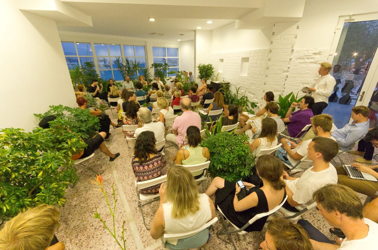 Public in Art Space Pythagorion's main hall seating in chairs among numerous cultivated plants. In front of the hall's window with a view to the sea, a group of speakers has gathered in circle and discusses.