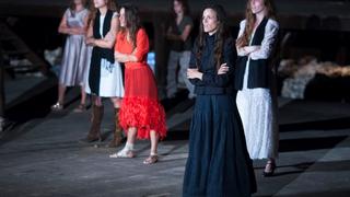 6 women in dresses standing on stage, perfoming the theatre play "Women of Troy", looking angrily with crossed arms