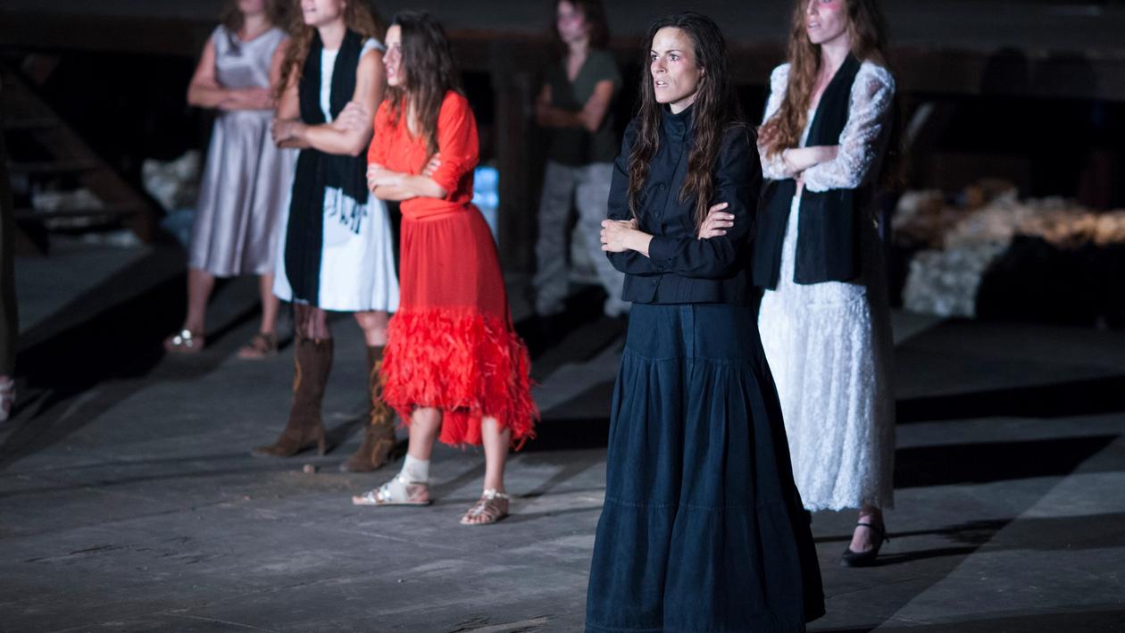 6 women in dresses standing on stage, perfoming the theatre play "Women of Troy", looking angrily with crossed arms