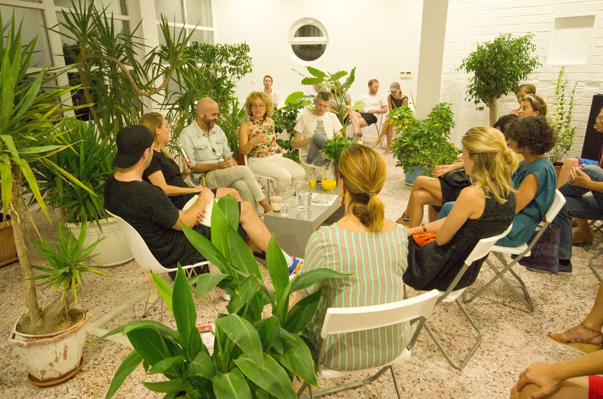 A group of speakers is seating in a circle interacting among numerous cultivated plants. One of them is holding and talking to the microphone. Behind them is Art Space Pythagorion hall's window with a view to the sea.  It is dark outside.