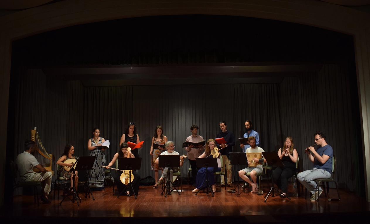 A group of people of different ages on stage arranged in two rows. In the front row 8 people are seating, each one playing a baroque instrument, i.e the flute, the lute, the recorder etc. Six people standing behind them are holding envelopes and they are singing. 