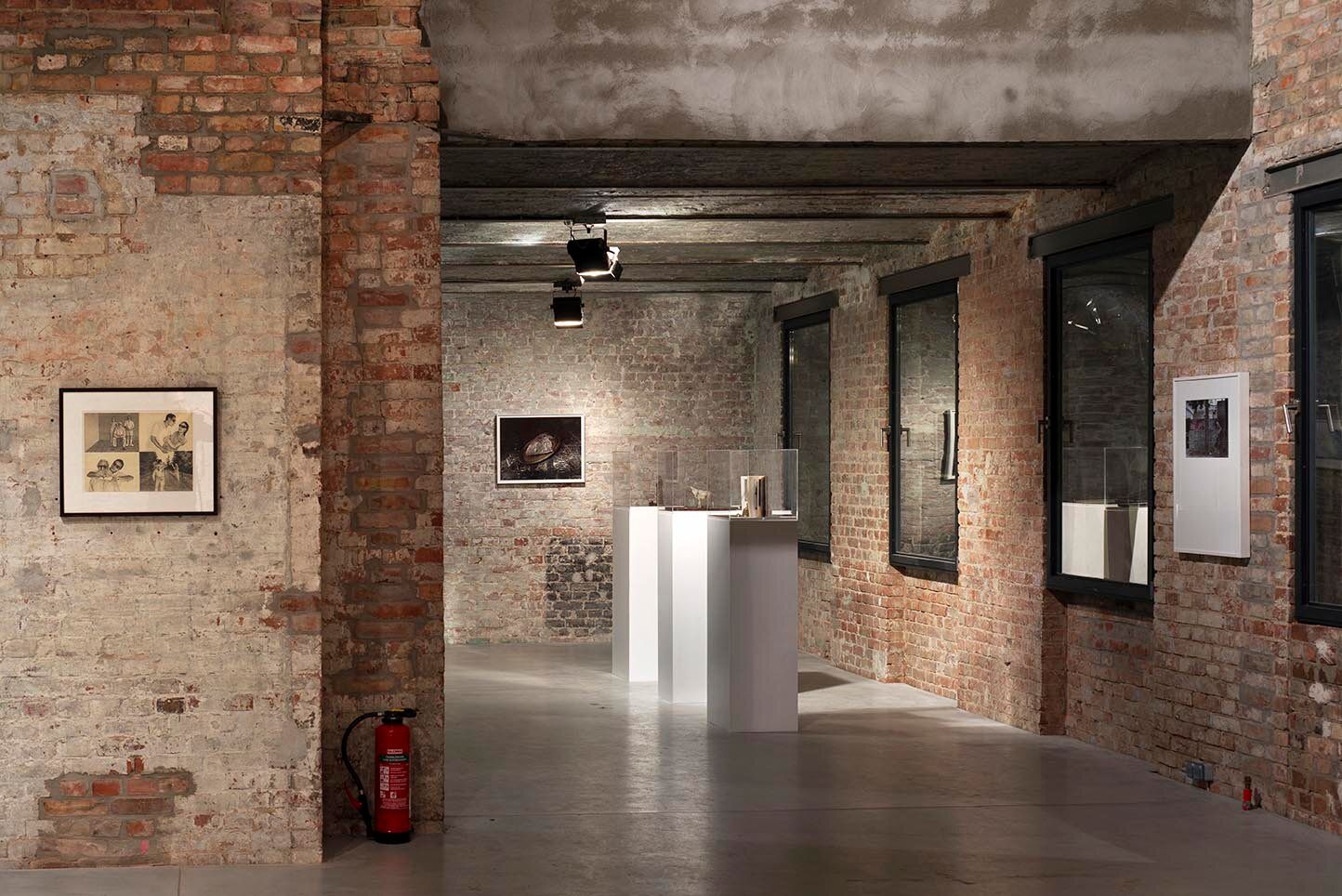 Interior of the exhibition space with walls made out of bricks and cement beams. Frames are hang on the wall and in the middle of the room are well-lit white showcase plints.