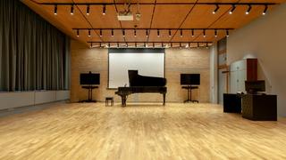 The distance learning studio is a room within the Athens Conservatoire building with wooden flooring and ceiling. On a brick wall there are two monitors between a blank projection screen. A grand piano, with its lid raised open is in front of the screen. To the left, there is a desk with a computer next to a standing cabinet.