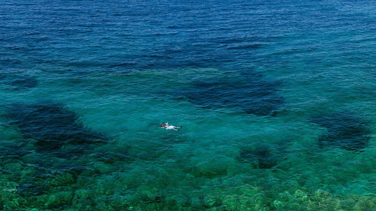 The blue sea photographed from above. In the bottom of the picture a man dressed in the greek tradiotional army soldier "tsolias" uniform and floates on the surface of the sea.