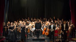 An orchestra with 40-50 musicians onstage bowing to the audience. In front of the orchestra are the singers and the conductor