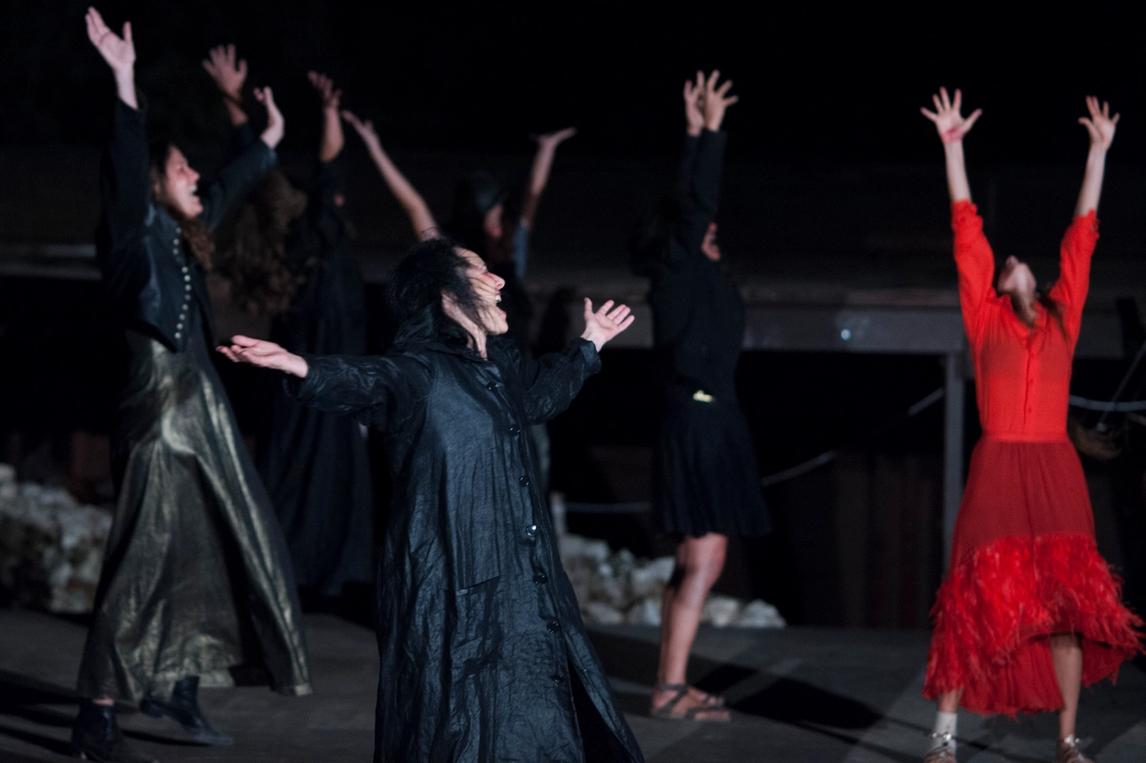 6 women, 5 in black one in red, reaching their arms to the sky