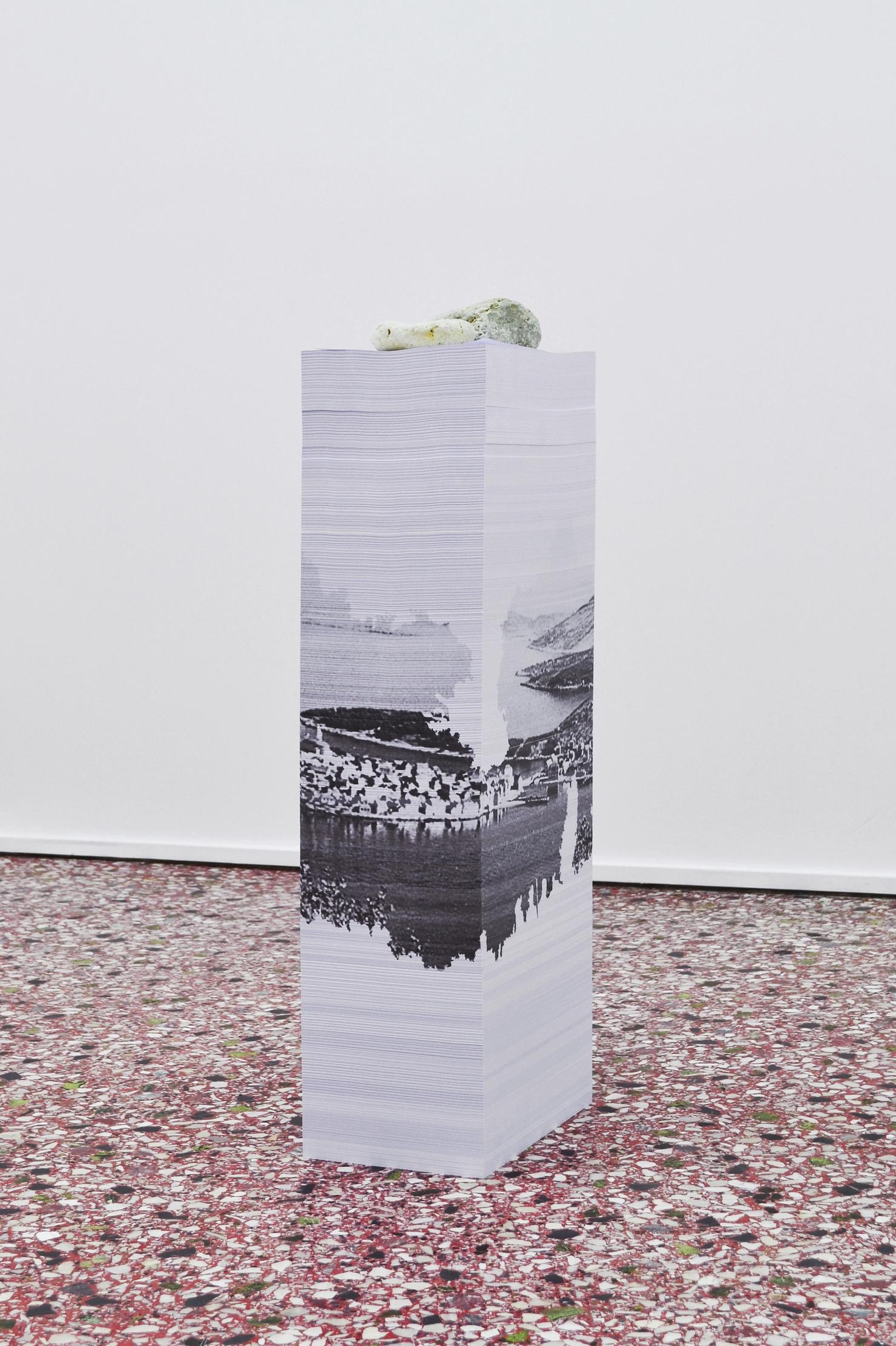 A single paper-stack sculpture comprised of A4 sheets of paper piled into columns in a red terrazzo floor and surrounded by white walls. A black and white landscape image is formed on the lateral sides of the stack.