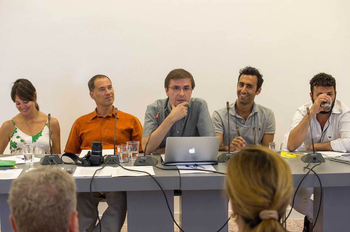 A group of speakers is seating at a grey table. A man in the middle is talking to the microphone in front of him while others are smiling.